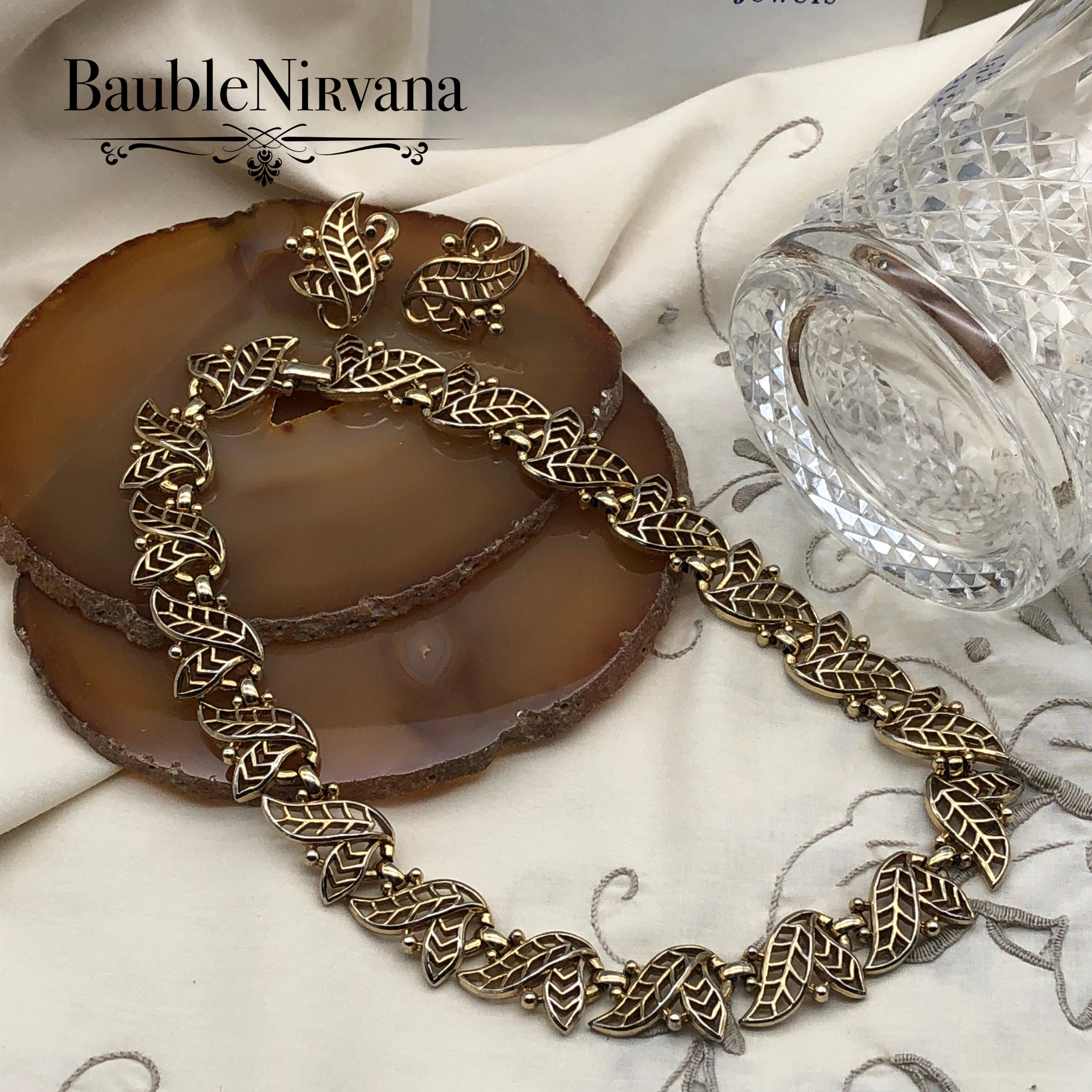 Gold leaf necklace and earrings on a decorative surface with 'BaubleNirvana' branding.