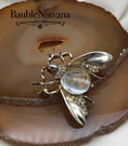 Load image into Gallery viewer, Gold bee-shaped brooch with a clear gemstone on a decorative stone background, featuring the brand 'BaubleNirvana'.
